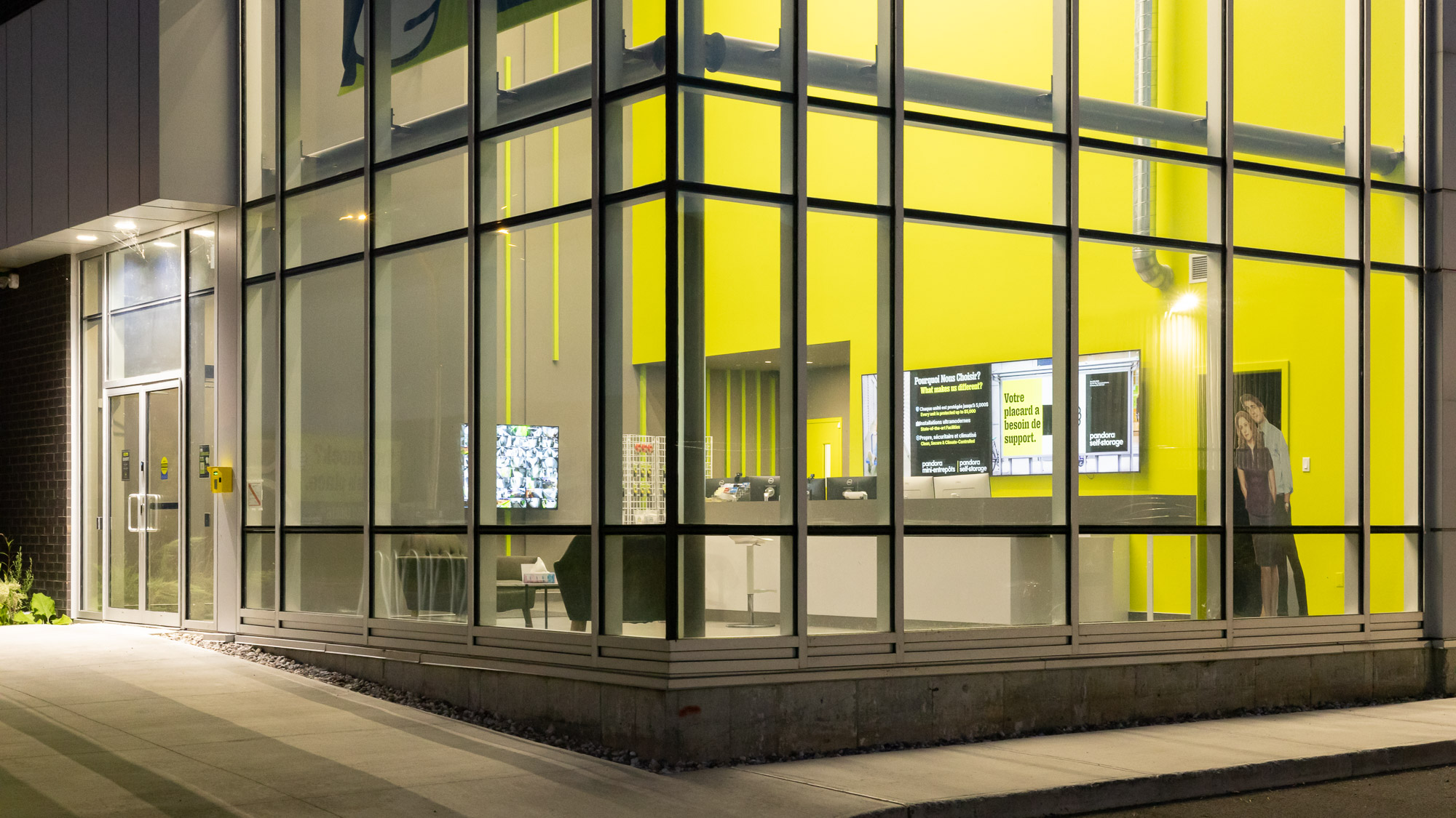 Entrée du bâtiment Pandora Mini-Entrepôts éclairée de nuit, avec façade vitrée et panneaux architecturaux jaunes.